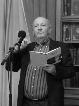 Derek Mahon at a poetry reading with a book in his left hand and a a microphone in the foreground